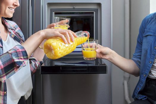 Close up of two womens hands pouring orange juice into glass from refrigerator in kitchen. People, food, drinks, diet, rest, communication, lifestyle concept