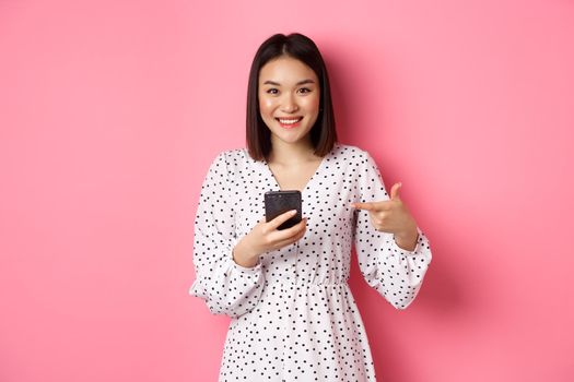 Beautiful young asian woman in romantic dress using smartphone, smiling and pointing at mobile phone, standing over pink background.