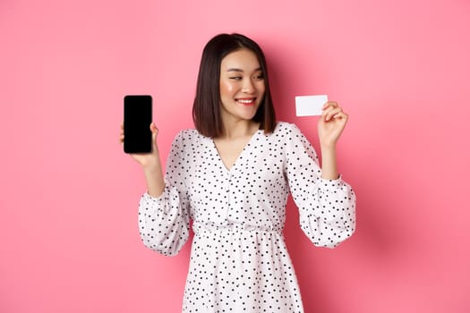 Happy young asian woman internet shopping, showing smartphone screen and looking satisfied at credit card, standing over pink background.
