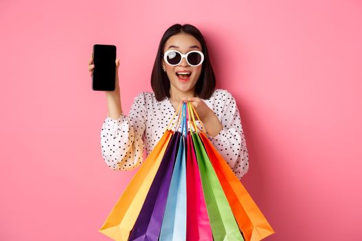 Attractive asian woman showing smartphone app and shopping bags, buying online via application, standing over pink background.