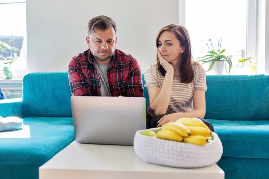 Serious middle aged couple looking at laptop screen at home in living room. Concentrated husband and wife listening looking into laptop, online information. Family, technology, lifestyle, 40s people