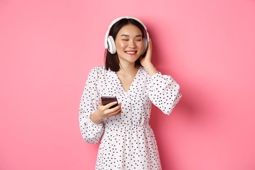 Romantic asian girl listening music in headphones, smiling with closed eyes, holding mobile phone, standing over pink background.