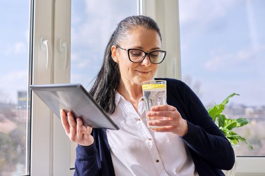 Adult woman drinks water with lemon, female in glasses with digital tablet, at home near window