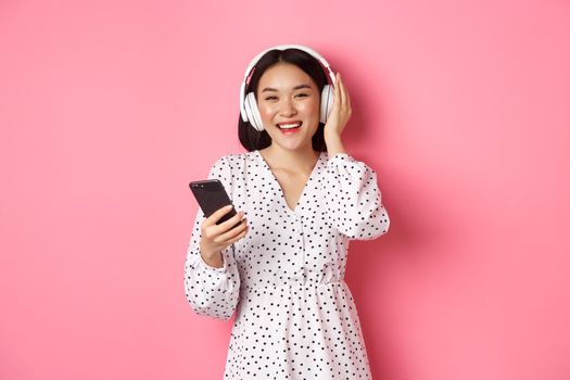 Beautiful asian woman listening music in headphones, using mobile phone, smiling happy at camera, standing over pink background.