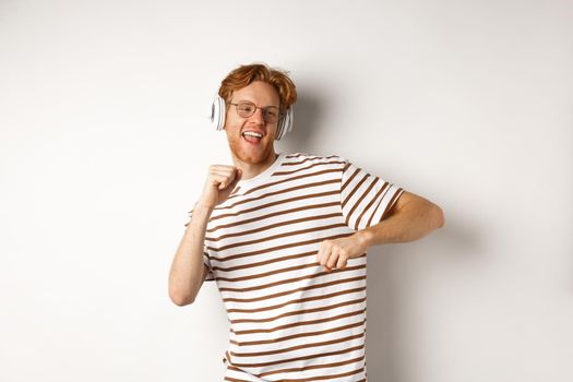 Technology concept. Happy redhead man listening music in headphones and dancing cheerful, standing in t-shirt against white background.