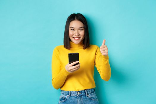 E-commerce and online shopping concept. Satisfied asian female client showing thumbs up and using smartphone, smiling pleased at camera, blue background.