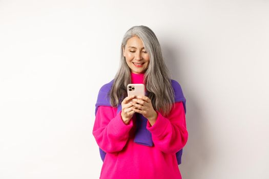 Beautiful asian senior woman in pink stylish sweater using smartphone. Old lady reading on mobile phone and smiling, standing over white background.