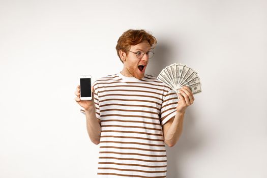 Amazed redhead man showing smartphone app on blank screen and money, winning prize cash online, standing over white background.