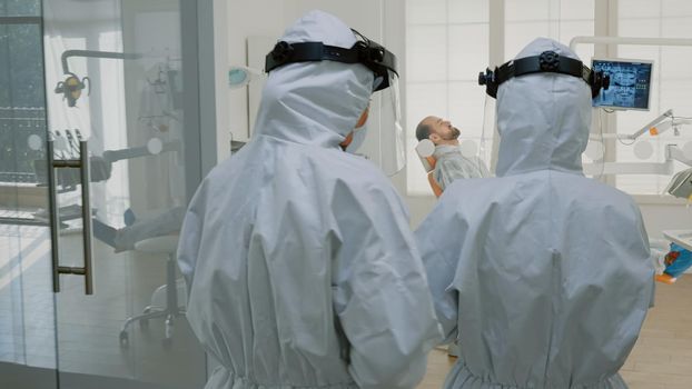 Orthodontist with ppe suit preparing for patient consultation using dental equipment and tools in oral cabinet. Dentist talking to assistant before starting teethcare examination