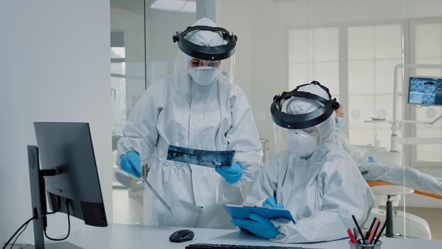 Orthodontist and assistant using x ray of teeth for oral care, wearing virus protection suits at clinic. Stomatologists looking at computer on desk and dental radiography in hand