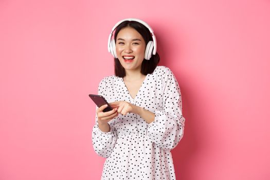 Beautiful asian woman listening music in headphones, using mobile phone, smiling happy at camera, standing over pink background.