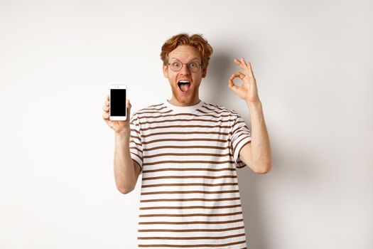 Technology and e-commerce concept. Young man with red hair showing okay sign and blank smartphone screen, praising awesome app, standing over white background.