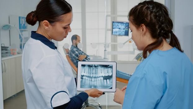 Dentist and nurse analyzing teeth radiography on tablet for modern oral care examination at dentistry clinic. Professional stomatological team using digital device for patient teethcare