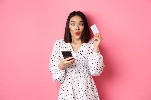 Attractive young asian woman order online, holding credit card and mobile phone, making internet purchase, standing happy over pink background.