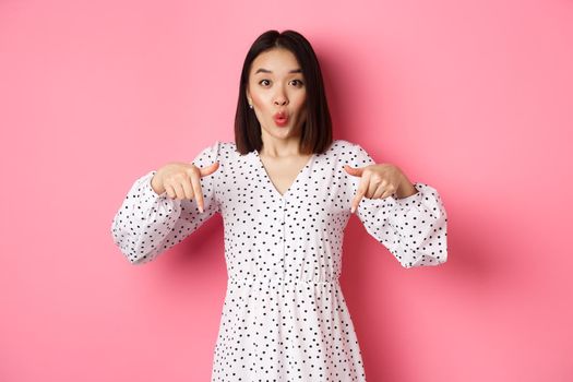 Impressed asian woman in dress pointing fingers down, look there gesture, check out discounts and sales, standing amazed over pink background.