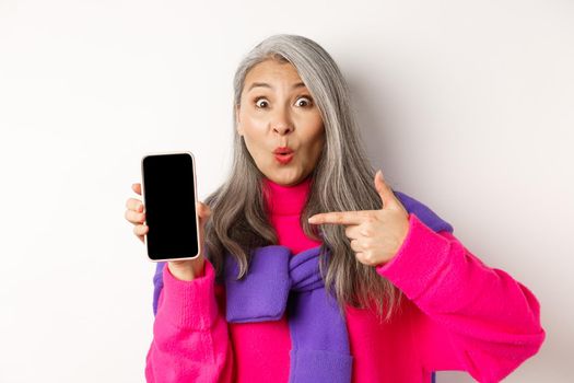 Online shopping. Beautiful asian grandmother smiling, pointing finger at smartphone blank screen, looking amazed, showing mobile application, standing over white background.