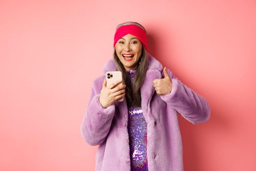 Online shopping and fashion concept. Happy asian grandmother checking out awesome promo, pointing finger at smartphone and looking amazed, pink background.