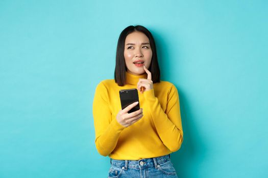 Pensive asian girl holding smartphone and thinking what to order online, standing over blue background.