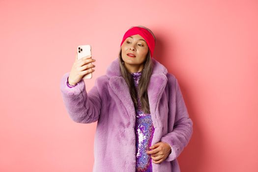 Fashion concept. Stylish asian senior female taking selfie on smartphone, posing in purple faux fur coat and party dress, standing over pink background.