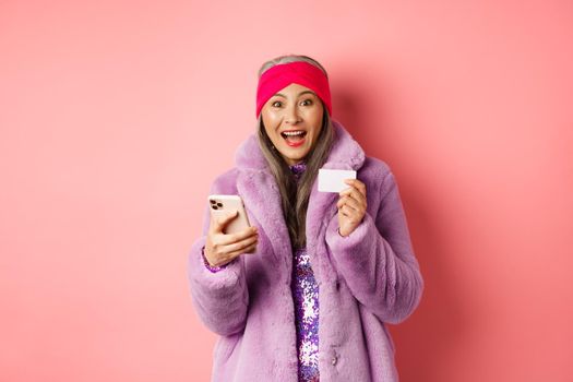 Online shopping and fashion concept. Happy asian female checking out internet promotions, holding credit card and smartphone, pink background.