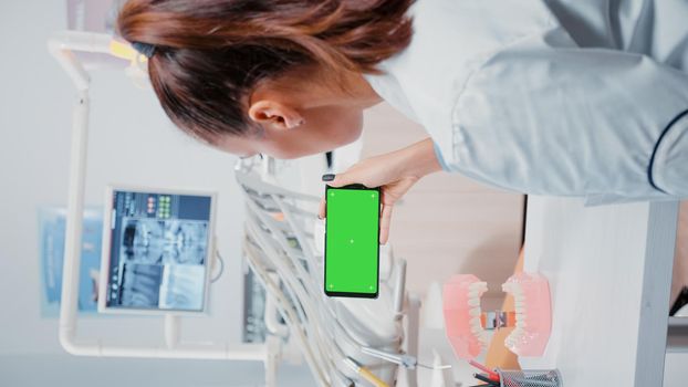 Vertical video: Specialist analyzing mobile phone with green screen for dental care. Dentist holding smartphone with chroma key on display for isolated background at stomatology clinic