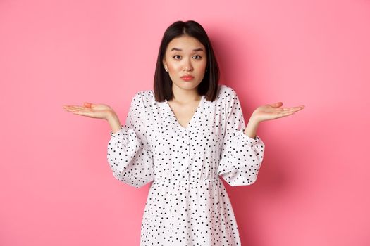 Indecisive cute asian woman shrugging, holding hands spread sideways on copy spaces, dont know, having doubts, standing over pink background.