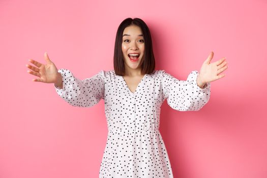 Happy asian girl stretching hands to camera, reaching for hug, smiling and wanting to hold something, standing over pink background in dress.