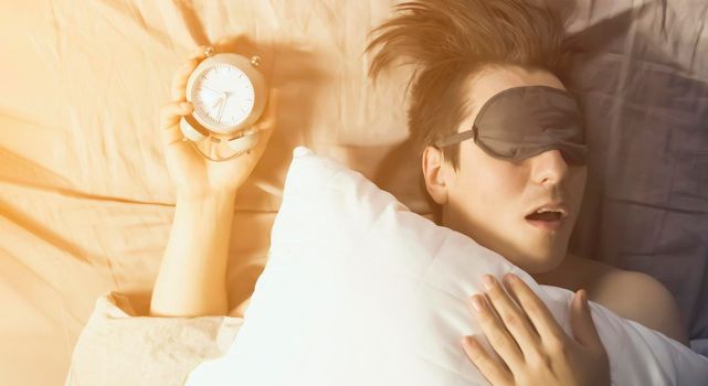A young funny man sleeps soundly early in the morning, hugs a pillow and holds a vintage alarm clock in his hands, the sleep mask has slipped from his eyes.