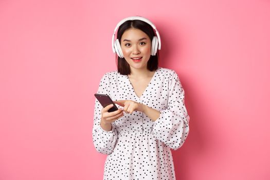 Beautiful asian woman listening music in headphones, using mobile phone, smiling happy at camera, standing over pink background.