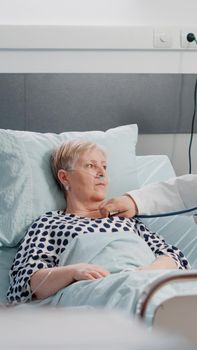 Medical team doing healthcare consultation for senior patient with illness. Doctor using stethoscope to examine sick woman with nasal oxygen tube in bed while nurse giving assistance