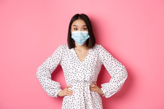 Covid-19, quarantine and lifestyle concept. Cute korean woman in dress and face mask using preventive measures from coronavirus, standing over pink background.