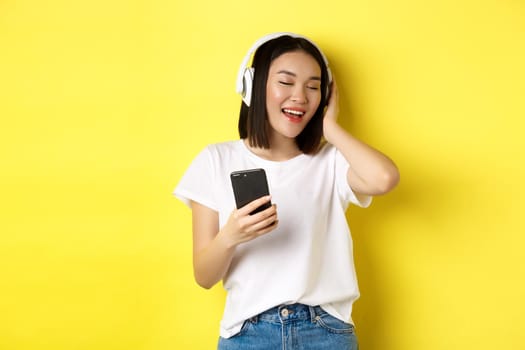 Cool asian girl singing along and listening music in wireless headphones, holding smartphone in hand, standing over yellow background.