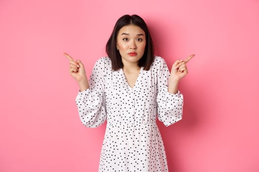 Confused asian woman hesitating, shrugging and pointing hands two sides, staring indecisive and need help with choice, standing over pink background.