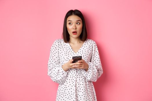 Online shopping. Confused asian girl holding smartphone, looking clueless at upper left corner, standing over pink background.