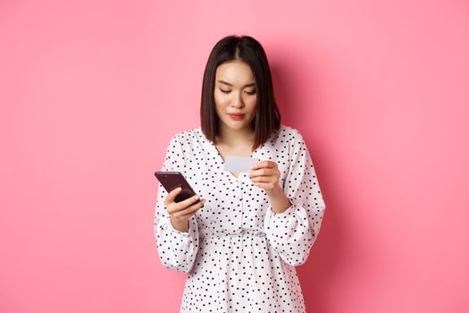 Beautiful asian woman enter credit card info in mobile phone app, shopping online, paying for order on smartphone, standing over pink background.