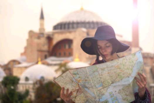 A young girl in a hat looks at a map of Istanbul on a sunny day. Beautiful ancient Hagia Sophia Mosque on the background. Traveling to Arab countries.