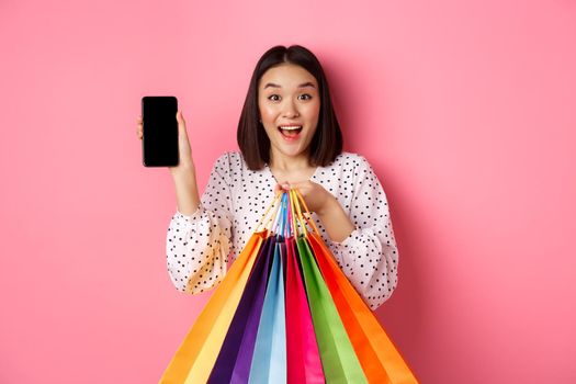 Attractive asian woman showing smartphone app and shopping bags, buying online via application, standing over pink background.