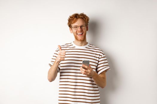 Technology and e-commerce concept. Satisfied male model with red hair, showing thumbs-up and holding smartphone, white background.