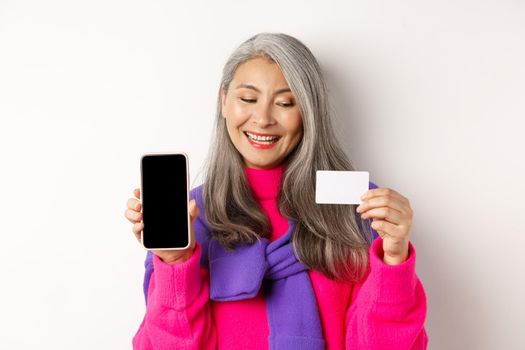 Online shopping. Closeup of fashionable old woman showing blank smartphone screen, looking pleased at plastic credit card, standing over white background.