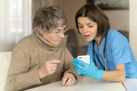A nurse in a medical suit takes care and explains to an elderly patient how to use applications on a smartphone. Grandmother 80 years old, does not understand how to communicate with a doctor online.
