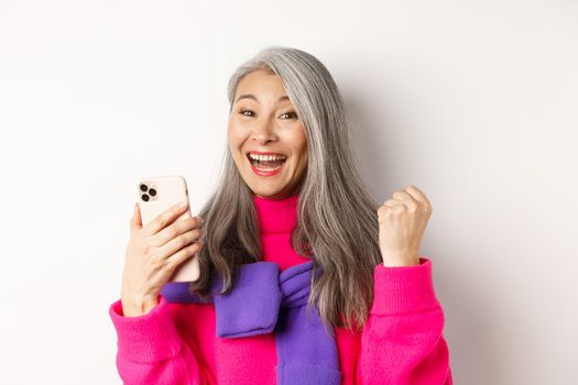 Online shopping. Close-up of cheerful asian elderly woman winning prize in internet, holding smartphone and making fist pump, standing over white background.