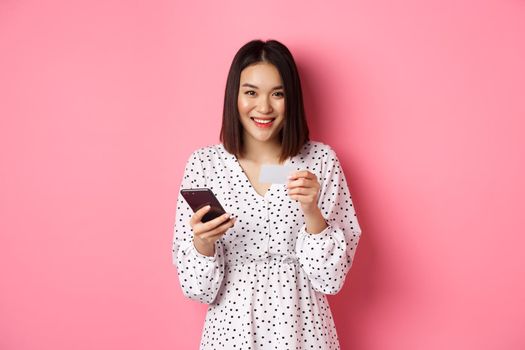 Beautiful asian woman enter credit card info in mobile phone app, shopping online, paying for order on smartphone, standing over pink background.