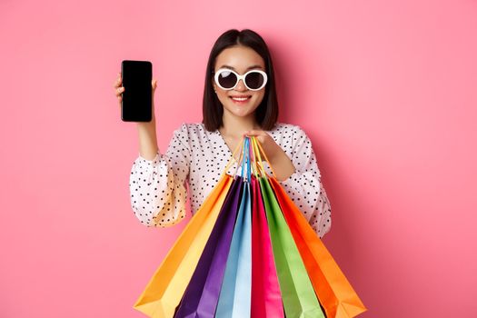 Attractive asian woman showing smartphone app and shopping bags, buying online via application, standing over pink background.