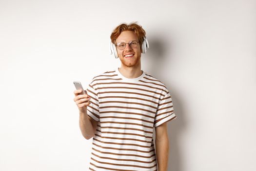 Technology concept. Young man with red hair and beard listening music in headphones and using smartphone, smiling at camera, white background.