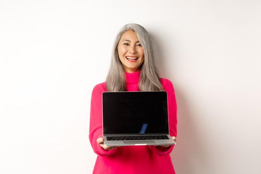 E-commerce concept. Beautiful asian senior businesswoman showing blank laptop screen and smiling, standing in pink sweater over white background.