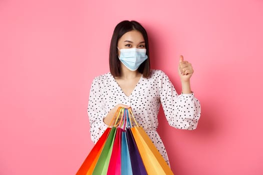 Covid-19, pandemic and lifestyle concept. Beautiful asian woman shopping in malls in medical mask, holding bags and showing thumb-up, standing over pink background.
