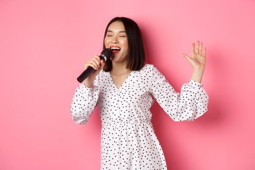 Happy asian woman dancing and singing in microphone, performing at karaoke, standing over pink background. Copy space