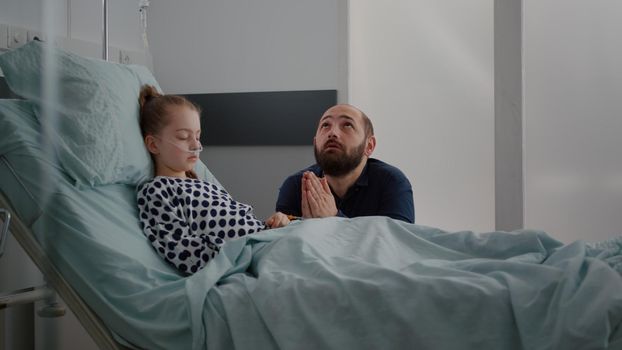 Sick girl daughter sleeping after surgery recovery while worried father praying for kid healthcare during medical consultation in hospital ward. Child wearing oxygen nasal tube resting in bed