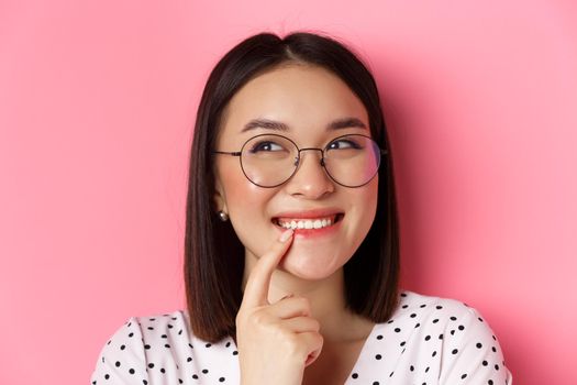 Headshot of cute asian woman in trendy glasses smiling, having an idea, thinking and looking at upper left corner, pink background.