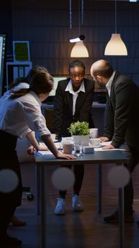 Diverse multi ethnic businesspeople teamwork lean on conference table having brainstorming session discussing company presentation. Coworkers overworking in meeting room late at night